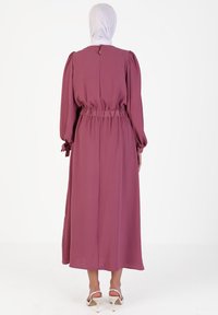 Long, gathered waist dress in dusty rose fabric, featuring long sleeves, a keyhole back, and a flowing skirt with ribbon accents at cuffs.