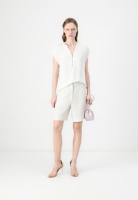 Top blanco de manga corta con cuello con cremallera, combinado con pantalones cortos beige claros. La modelo sostiene un pequeño bolso de mano lavanda. Sandalias nude con tiras.