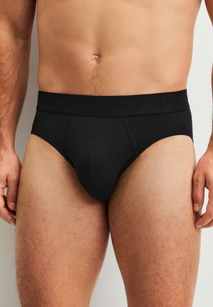 Man draagt zwarte slips met een gebrandmerkte tailleband, getoond van taille tot halverwege de dij tegen een effen achtergrond.