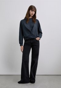 Donkergrijze sweatshirt met een uniek overslagontwerp, geribbelde zoom, gecombineerd met zwarte jeans met wijde pijpen. Haar valt in zachte golven. Soepele structuur.