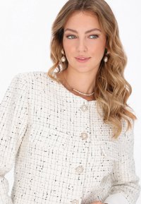 Chaqueta blanca texturizada con un patrón de cuadrícula, presenta botones en forma de perlas y bolsillos. La modelo lleva pendientes de perlas y collares.