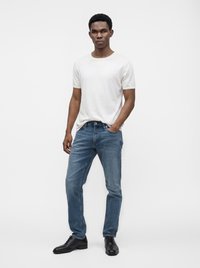 Un homme se tient devant un fond uni, vêtu d'un t-shirt blanc, d'un jean bleu et de chaussures habillées noires, une main dans la poche.