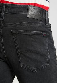 Jeans de denim negro con un parche de cuero etiquetado "TOMMY JEANS" en la cinturilla, que cuentan con bolsillos estándar y detalles de costura sutiles.