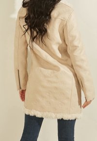 Abrigo beige con un patrón texturizado, puños con cremallera y forro peludo en el dobladillo. Mangas largas y diseño suelto de longitud media.