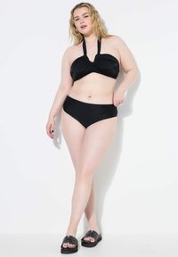 Set bikini nero con top bandeau torcido e spalline all'halterneck, accompagnato da slip a vita alta. Modella che indossa sandali neri.