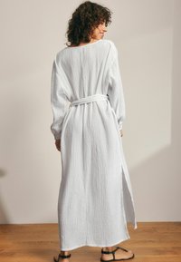 Robe longue blanche en tissu texturé, avec des manches longues, une ceinture à la taille et des fentes sur les côtés pour plus de liberté de mouvement.