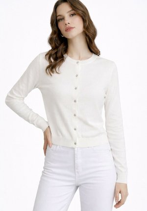 Jeune femme aux cheveux bruns ondulés portant un cardigan blanc boutonné et un pantalon blanc taille haute, posant avec une main sur la hanche.