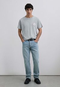 Camiseta de algodón gris con un parche de logotipo en forma de diamante, combinada con jeans de mezclilla azul claro y zapatos negros. Diseño de atuendo simple y casual.