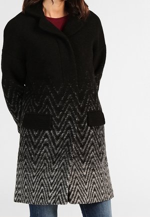 Femme portant un manteau dégradé du noir au gris avec un motif chevrons et deux poches avant noires, associé à un jean bleu et un haut rouge foncé.