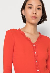 Top de manga longa riscado em vermelho com decote em V decorado com renda e botões brancos na frente. Textura suave, design ajustado.