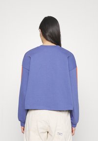 Una donna con lunghi capelli scuri indossa una felpa a maniche lunghe blu con strisce arancioni sulle maniche e pantaloni leggeri beige con un'etichetta di marca visibile.