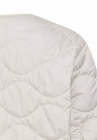 Veste matelassée beige clair avec un motif ondulé ; fabriquée en tissu lisse avec une texture douce et un détail de couture proéminente.