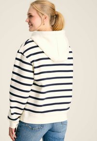 Pruhovaná mikina v krémovej a námorníckej modrej, so skladacou kapucňou, ribbovanými manžetami a spodným lemom. Mäkká tkanina s voľným strihom, viditeľné švy a ležérny dizajn.