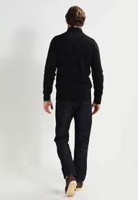 Maglione nero a collo alto, abbinato a jeans blu scuro. Scarpe marrone chiaro con suola texturizzata, viste da dietro.
