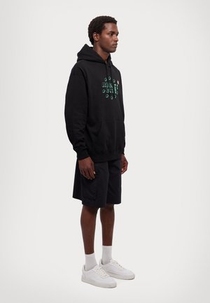 Black hooded sweatshirt features a front pocket and green graphic design. Paired with black shorts and white sneakers, worn with white socks.