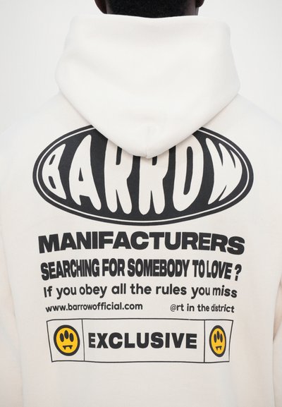 Vit hoodie med en stor svart grafisk tryck på baksidan med text och ett smiley-ansikte, som visar en avslappnad design och mjuk textur.