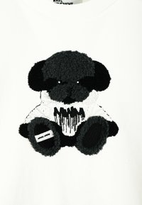 Aplicación de un oso de peluche negro y gris sobre una prenda blanca, con pelo texturizado, una camiseta con diseño de goteo negro y acentos de tela en contraste.
