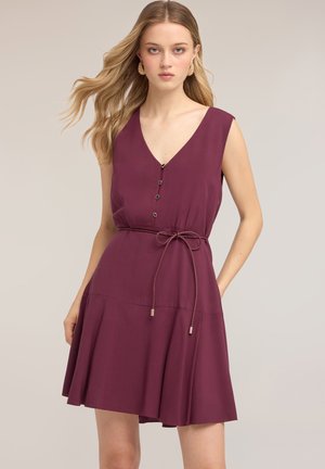 Femme portant une robe prune sans manches avec boutons et ceinture à la taille, se tenant devant un fond clair uni.