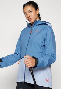 Chaqueta impermeable azul claro con capucha. Presenta acentos en azul más oscuro, cierre de cremallera y puños ajustables. Evoca una preparación para actividades al aire libre.