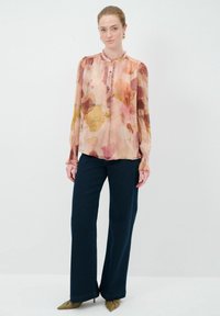 Blumenbluse mit einem sanften rosa Grundton, die orangefarbene und kastanienbraune Blättermuster zeigt, eine Knopfleiste hat und gesammelte Ärmel besitzt. Kombiniert mit dunkelblauen Jeans.