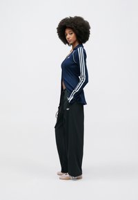 Navy krāsu sporta jaka ar baltām svītrām, atvērta priekšpuse, kopā ar vaļīgām melnām biksēm. Jakā un biksēs redzams Adidas logotips.