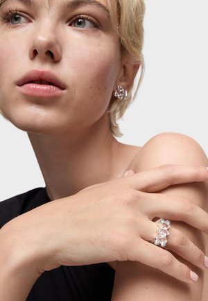 Anillo de plata con múltiples piedras claras, con un diseño floral. La modelo lleva pendientes a juego y una blusa sin mangas negra.
