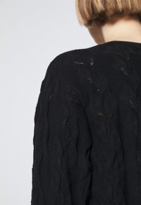 Taglio di capelli bob corto visibile da dietro, indossando un maglione lavorato a maglia nero con un motivo a foglia testurizzata su uno sfondo chiaro e semplice.