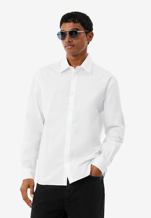 Joven con gafas de sol negras, una camisa blanca de manga larga abotonada y pantalones negros, con una mano en el bolsillo.
