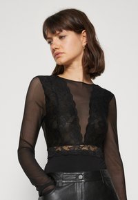 Bodysuit de encaje negro con mangas largas transparentes, escote en V profundo y detalles de encaje con borde en la cintura, combinado con pantalones de cuero negros.