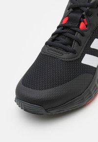 Chaussure de sport noire avec une tige en tissu texturé, dotée de rayures blanches et d'accents rouges. Bout arrondi et semelle en caoutchouc pour une meilleure traction.