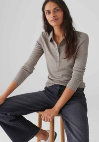 Hellgraues, langärmliges Polohemd mit Knopfleiste, kombiniert mit dunklen Jeans. Das Model sitzt auf einem Holzstuhl.