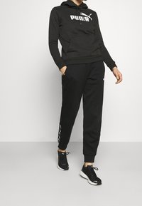 Felpa nera con logo Puma bianco, abbinata a pantaloni della tuta neri con dettagli laterali. Progettato per il comfort, adatto per un abbigliamento casual.