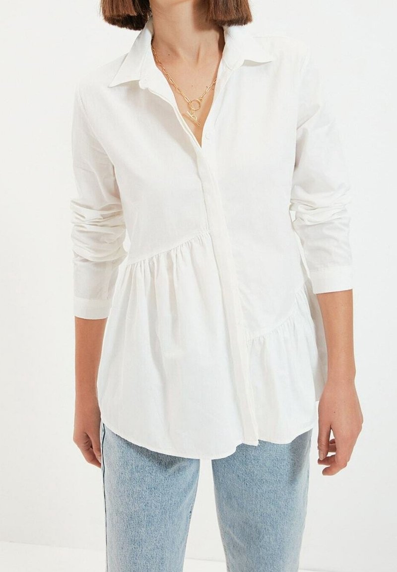 Chemise blanche boutonnée en tissu léger, avec un ourlet plissé, des manches longues et un col classique. Associée à un jean bleu clair.