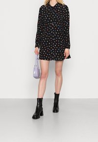 Czarna sukienka z kolorowymi, liściastymi wzorami, długimi rękawami i kołnierzykiem; zestawiona z czarnymi botkami na obcasie i małą lawendową torebką.