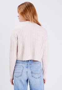 Helle beige, cropped Strickjacke mit vertikalen Rippungen, rundem Halsausschnitt und langen Ärmeln, kombiniert mit hochtaillierten, hellen Jeans.