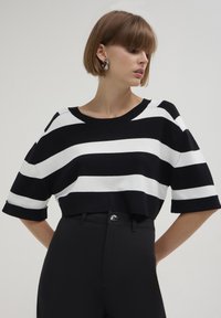 Gestreifter cropped Sweater in Schwarz-Weiß mit weitem Ausschnitt und kurzen Ärmeln, aus einem gestrickten Stoff gefertigt. Kombiniert mit hochgeschnittenen schwarzen Hosen.