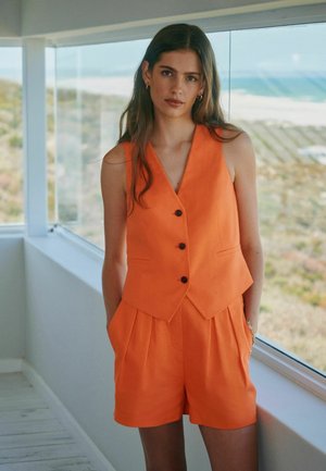 Frau mit langen braunen Haaren, die eine orange ärmellose Weste und passende Shorts trägt, steht drinnen an einem großen Fenster mit Blick auf die Natur draußen.