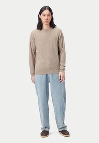 Beige texturerad tröja med långa ärmar och rund hals, kombinerad med ljusblå jeans med lös passform och bruna flätade loafers. Enfärgad bakgrund.