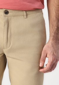 Beige cotton chinos with a smooth texture, featuring a standard button closure and side pockets, partially shown with a hand touching the fabric.