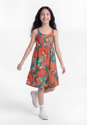 Mosolygó lány színes virágos ujjatlan ruhában sétál, fehér tornacipőt és virágmintás fehér zoknit visel.