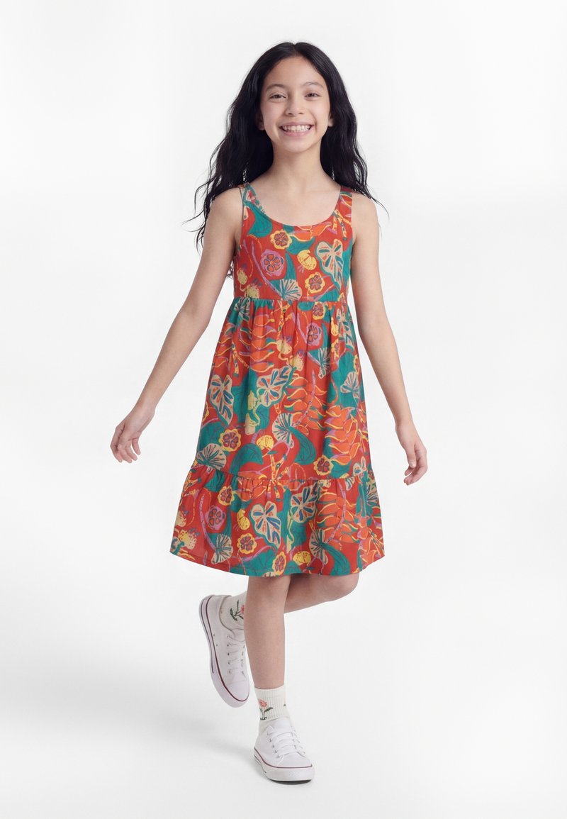 Χαμογελαστή κοπέλα σε πολύχρωμο, χωρίς μανίκια, φόρεμα με λουλούδια, περπατώντας, φοράει λευκά αθλητικά παπούτσια και λευκές κάλτσες με λουλουδάτα σχέδια.