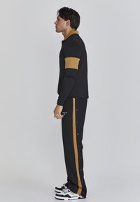 Survêtement noir avec des accents dorés ; le haut à manches longues présente un col doré et des manches dorées, tandis que le pantalon est doté de boutons-pression sur le côté.