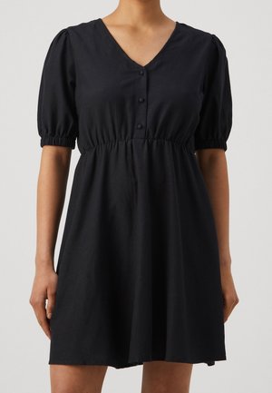 Mujer vistiendo un vestido negro de manga corta con escote en V, tres botones en el frente, cintura fruncida y puños elásticos en las mangas.