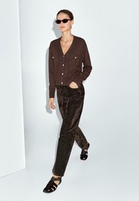 Cardigan lavorato a maglia marrone con bottoni dorati, abbinato a pantaloni in velluto marrone scuro e sandali neri con cinturini. La modella cammina su uno sfondo semplice.