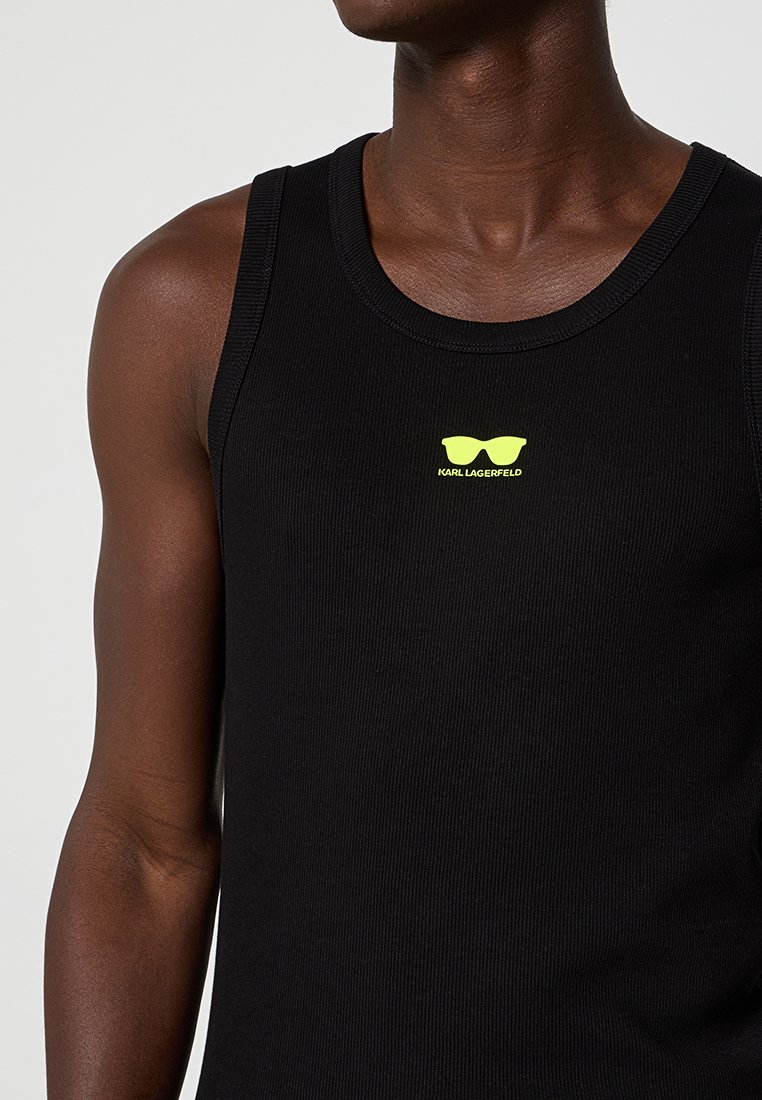 Top sin mangas de canalé negro llevado por una persona de piel oscura, con un pequeño logo de gafas de sol en neón amarillo y el texto "KARL LAGERFELD" en el pecho.