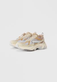 Beige und graue Sportschuhe mit Mesh-Einsätzen, strukturierten Details und runden Sohlen. Sie verfügen über elastische Schnürsenkel und eine kleine Zuglasche.