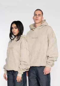 Helle beigefarbene Hoodies mit weicher Textur, ausgestattet mit einer Kängurutasche und einem zentralen Logo. Kombiniert mit dunklen Jeans.