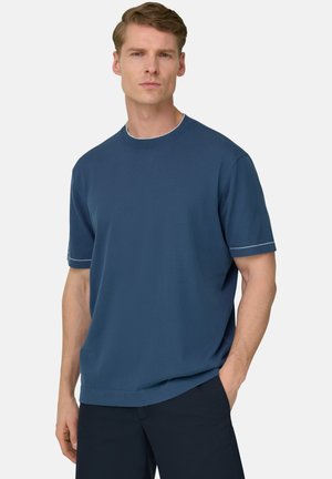 Mann mit kurzen hellbraunen Haaren, der ein blaues kurzärmeliges Hemd und dunkle Hosen trägt, steht mit einer Hand in der Tasche.