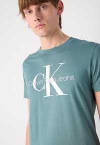 Tamsiai mėlynas medvilnės marškinėliai su dideliu baltu logotipu, kuriame užrašyta "Calvin Klein Jeans". Paprastas apvalios iškirptės dizainas, trumpomis rankovėmis.