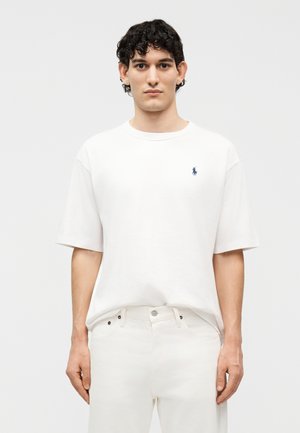 Νεαρός άνδρας με κοντά σγουρά μαλλιά, φοράει λευκό κοντομάνικο πουκάμισο με ένα μικρό κεντημένο λογότυπο και λευκό παντελόνι, στέκεται μπροστά σε απλό φόντο.